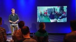 Person holder præsentation på en scene foran publikum med et projiceret billede på skærmen.