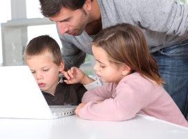 Father assisting two children while they use a laptop at a table.
