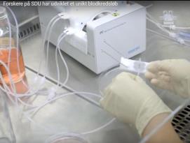 Person med handsker arbejder med en hvid laboratorieenhed og flere gennemsigtige slanger på et laboratoriebord.