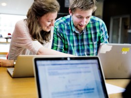 Two students collaborating at a table with laptops, with a blurred laptop in the foreground.