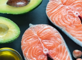 Two raw salmon steaks beside halved avocados, olive oil, and hazelnuts on a dark background.