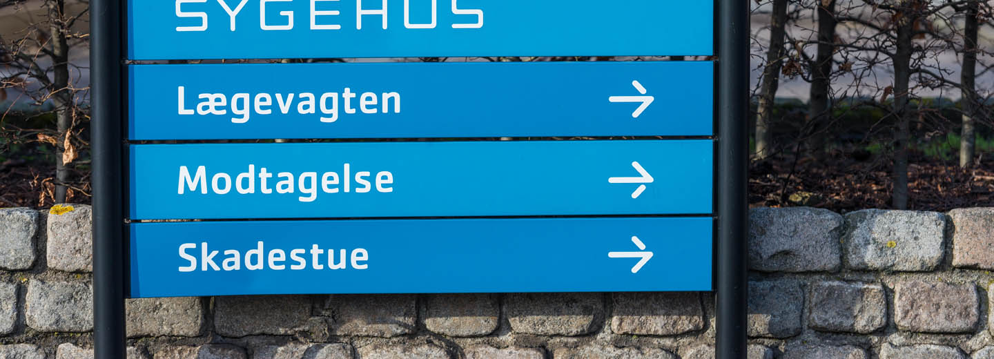 Blåt skilt ved et sygehus med teksten “SYGEHUS”, “Lægevagten”, “Modtagelse” og “Skadestue” samt højrepile.