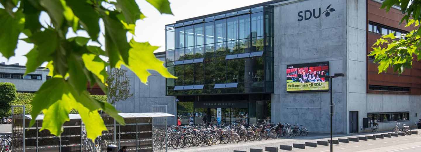Universitetsbygning med glasfacade, cykelparkering foran og grønne blade i forgrunden. På facaden står "SDU", og en skærm viser "Velkommen til SDU".