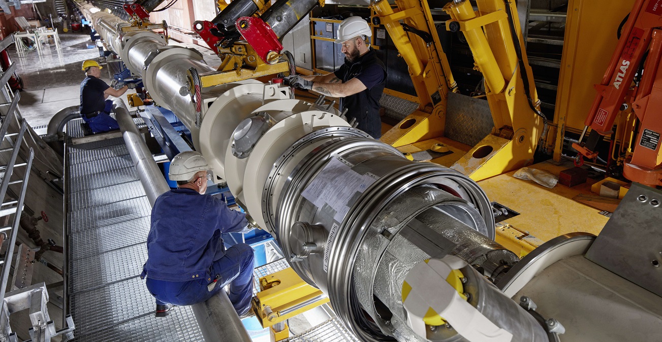 Tre teknikere udfører arbejde på en stor cylindrisk maskine i en industrifacilitet med gule løftearme og synlig tekst "ATLAS" på en orange arm.