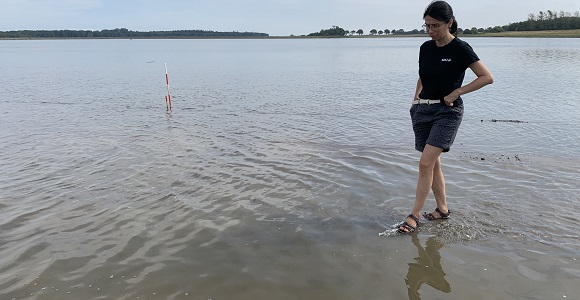 Person walking in shallow lagoon water near a marker pole with a distant shoreline behind.
