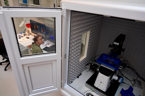 Person ved computerarbejdsstation ved siden af en åben akustisk testkabine med laboratorieudstyr indeni.