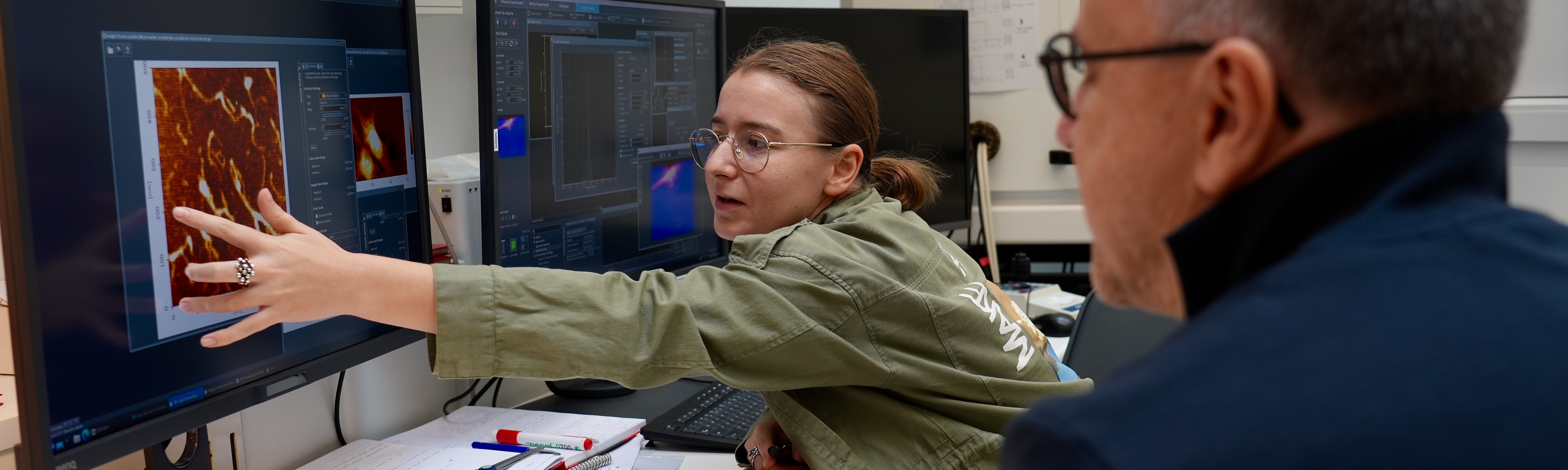 Person i laboratorie peger på et mikroskopisk billede på en computerskærm, mens en anden person observerer.