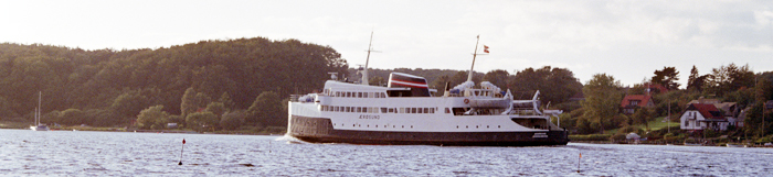 White ferry on calm water near a wooded shoreline and houses in the background.
