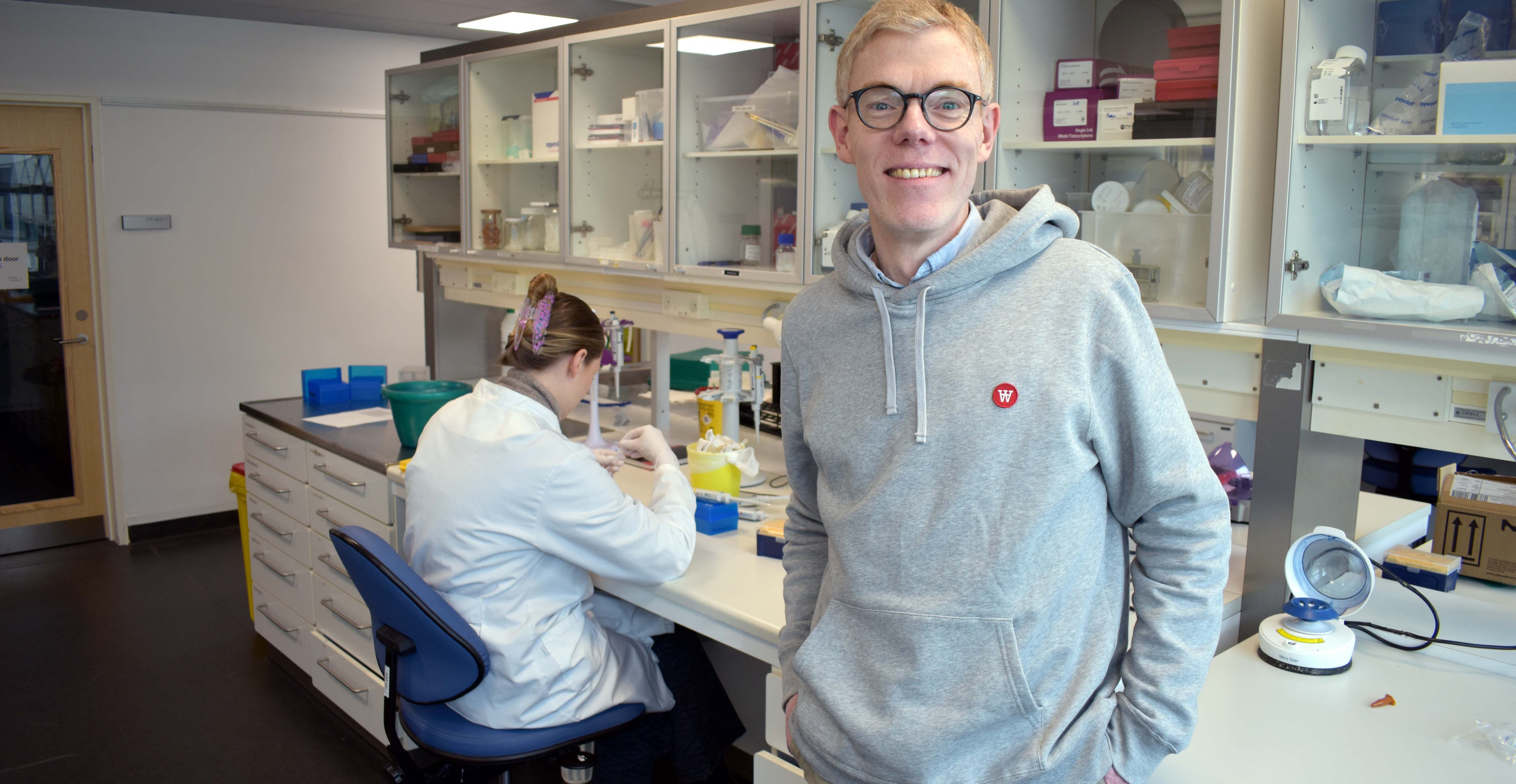 Kim Ravnskjær i et laboratorium med videnskabeligt udstyr og en person i kittel, der arbejder ved et skrivebord.