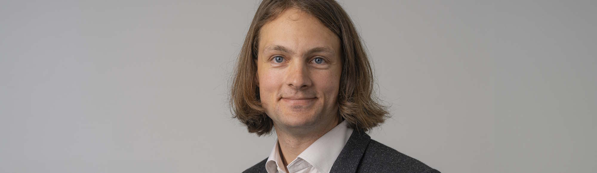 Beskåret portræt af en smilende mand med skulderlangt hår i skjorte og blazer mod grå baggrund.