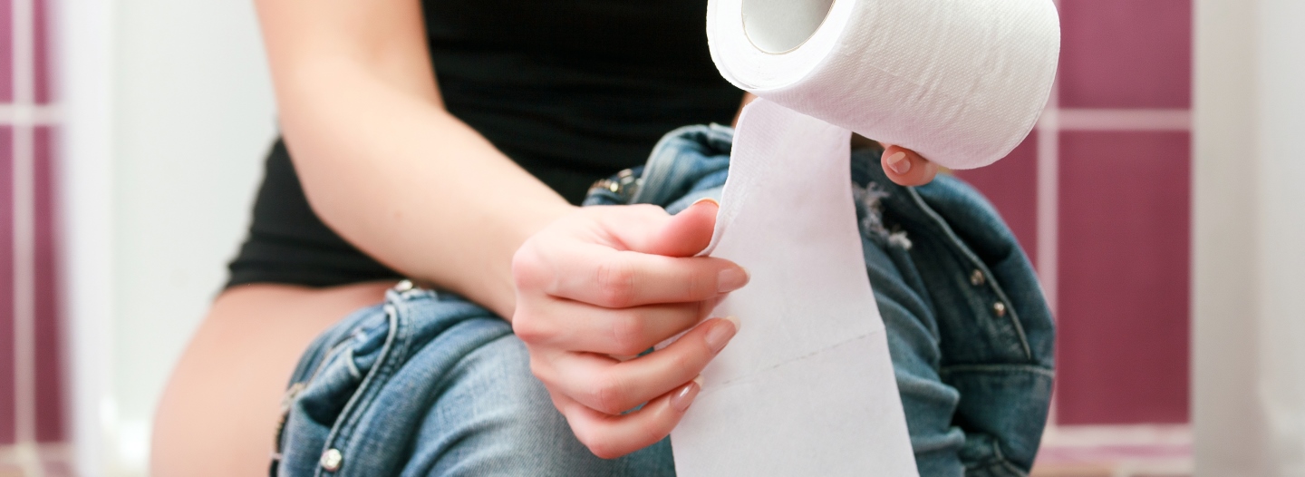 Kvinde sidder på toilet med toiletpapir og nedtrukne jeans i et badeværelse.