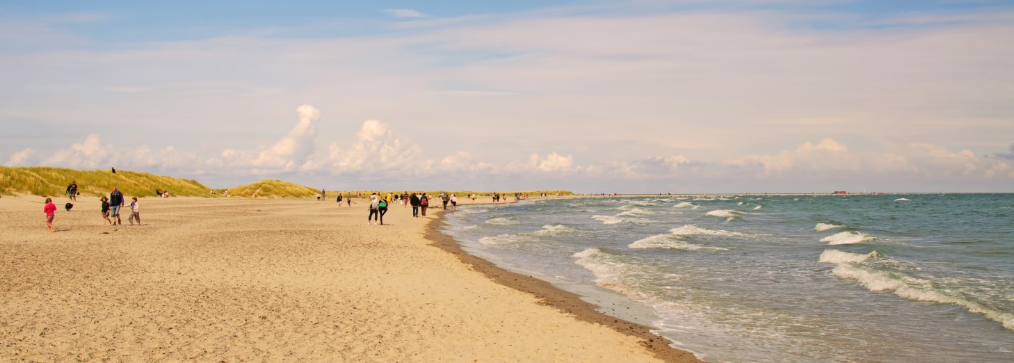 Bred sandstrand med klitter, spredte mennesker langs kysten og små bølger ved vandkanten under en let skyet himmel.