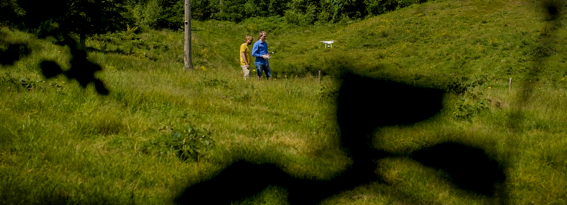 To personer i et grønt landskab med en hvid drone i luften og mørke, uskarpe former i forgrunden.