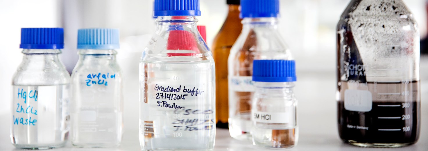 Glasflasker med blå låg og kemikalier på et laboratoriebord med etiketter som "HgCl2 ZnCl2 waste", "avfald zink", "Gradient buff 27.4.2015 J.Fowler" og "3M HCl".