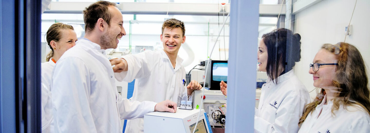 Fem forskere i hvide kitler samtaler og smiler i et laboratorium med teknisk udstyr.