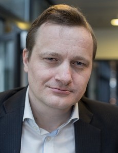Portræt af en mand i mørkt jakkesæt og hvid skjorte med sløret indendørs baggrund.