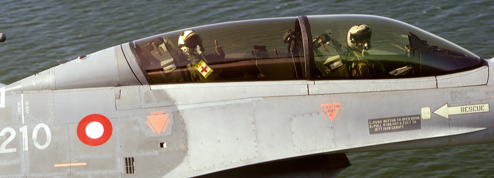 Nærbillede af et F-16-flys cockpit med to personer i flyvehjelme og dansk rundel på siden.