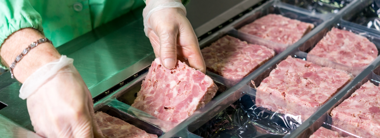 Hænder med handsker pakker forarbejdet kød i plastbakker på en produktionslinje.
