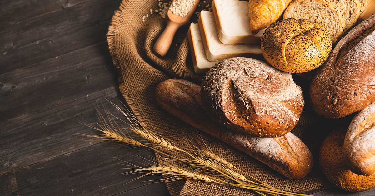 Blandet udvalg af friskbagt brød og bagværk, herunder fuldkornsbrød, baguettes, croissanter og franskbrød, anrettet på rustikt træbord med hvedekerner og stofdug.