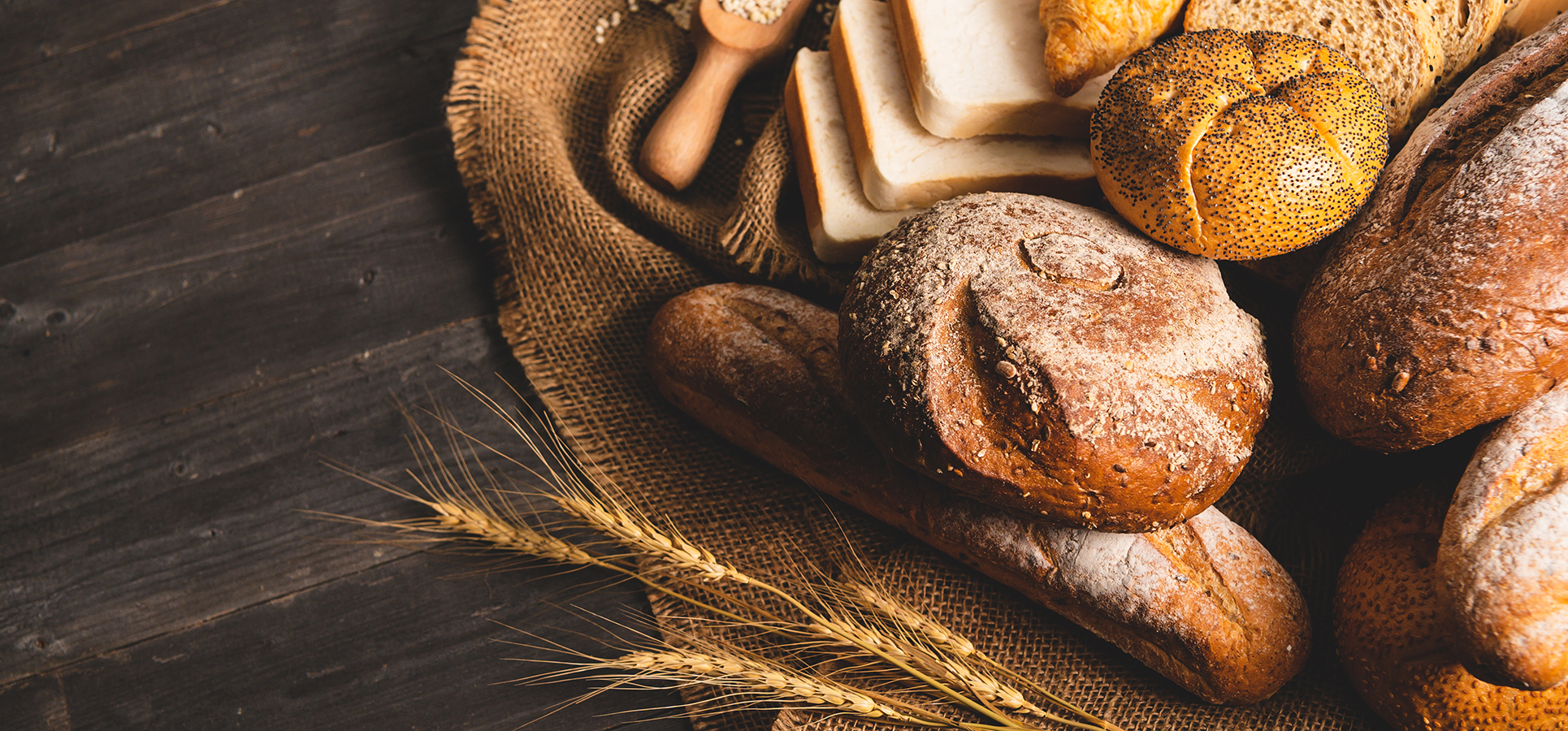 Blandet udvalg af friskbagt brød og bagværk, herunder fuldkornsbrød, baguettes, croissanter og franskbrød, anrettet på rustikt træbord med hvedekerner og stofdug.