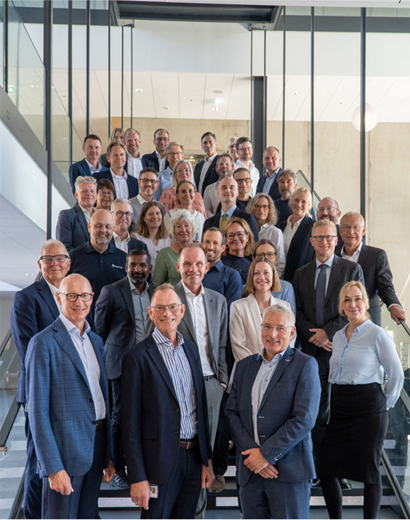 Stor gruppe professionelt klædte personer poserer til et gruppefoto på en trappe i en moderne kontorbygning.