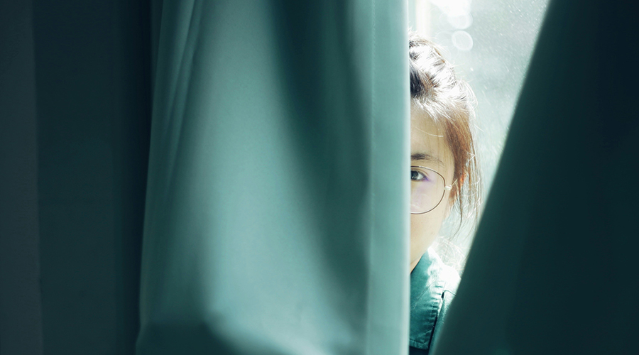 Kvinde kigger nysgerrigt gennem et gardin ved et solbeskinnet vindue. Lyset skaber en stemningsfuld kontrast mellem skygger og det oplyste ansigt.