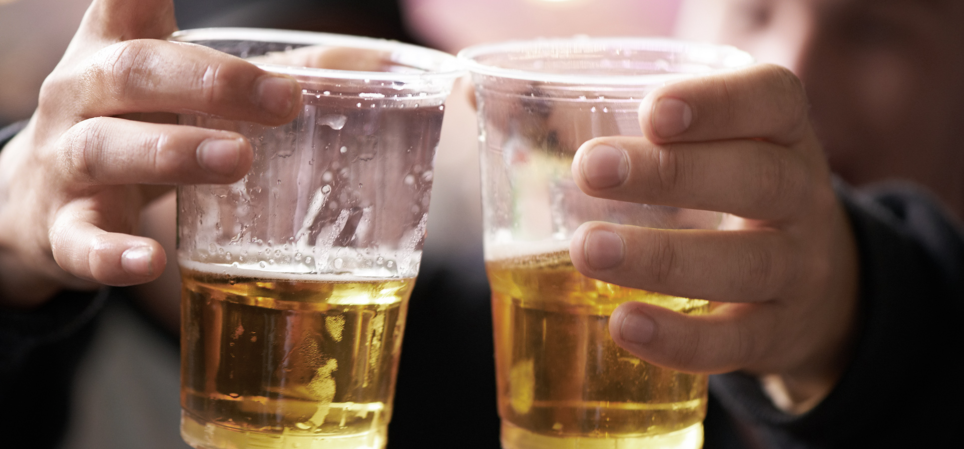 Nærbillede af to personer, der skåler med plastikkrus fyldt med øl ved en fest eller social begivenhed. Gyldent øl, feststemning og sløret baggrund med varme lys.