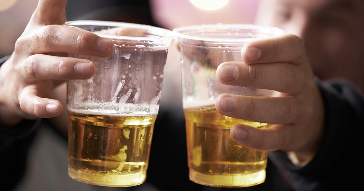 Nærbillede af to personer, der skåler med plastikkrus fyldt med øl ved en fest eller social begivenhed. Gyldent øl, feststemning og sløret baggrund med varme lys.