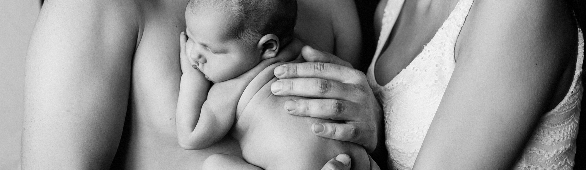 Sort-hvidt familiefoto af en nyfødt baby, der sover trygt i sin fars arme, mens moren kærligt holder om dem begge.