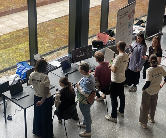 Besøgende står ved en informationsstand indendørs og taler med formidlere under et videnskabsarrangement, med computere og skærme på bordet.