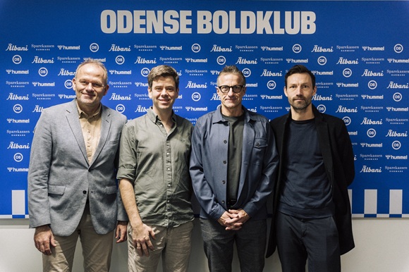 Fire mænd står foran en blå væg med logoer fra sponsorer og teksten "Odense Boldklub". De poserer til et officielt foto, sandsynligvis i forbindelse med en pressemeddelelse eller præsentation hos den danske fodboldklub OB.