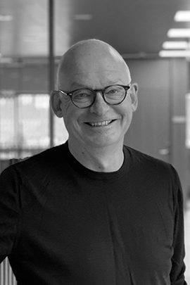 Sort-hvidt portrætfoto af en smilende, skaldet mand med briller og sort T-shirt i et moderne kontormiljø.
