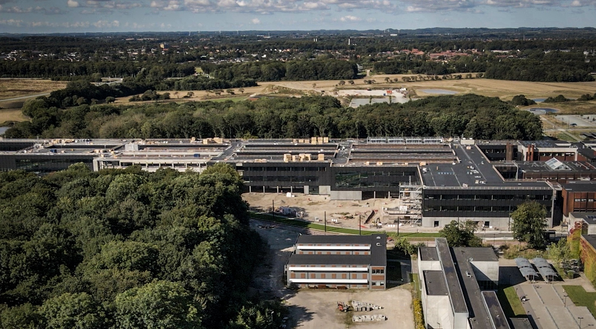 Luftfoto af et stort bygningskompleks med skov i forgrunden og marker samt byområde i baggrunden.