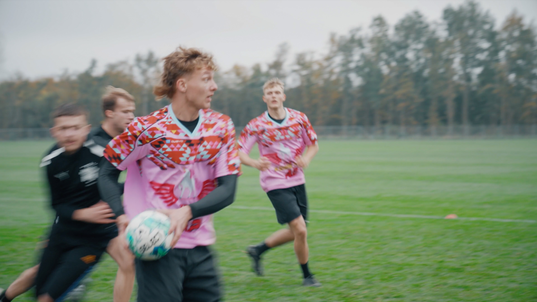 Unge mænd spiller rugby på en græsmark, iført farverige pink og røde trøjer. En spiller i forgrunden holder bolden og kigger bagud, mens med- og modspillere løber omkring ham. Stemningen er intens, og spillerne er i bevægelse midt i kampen.