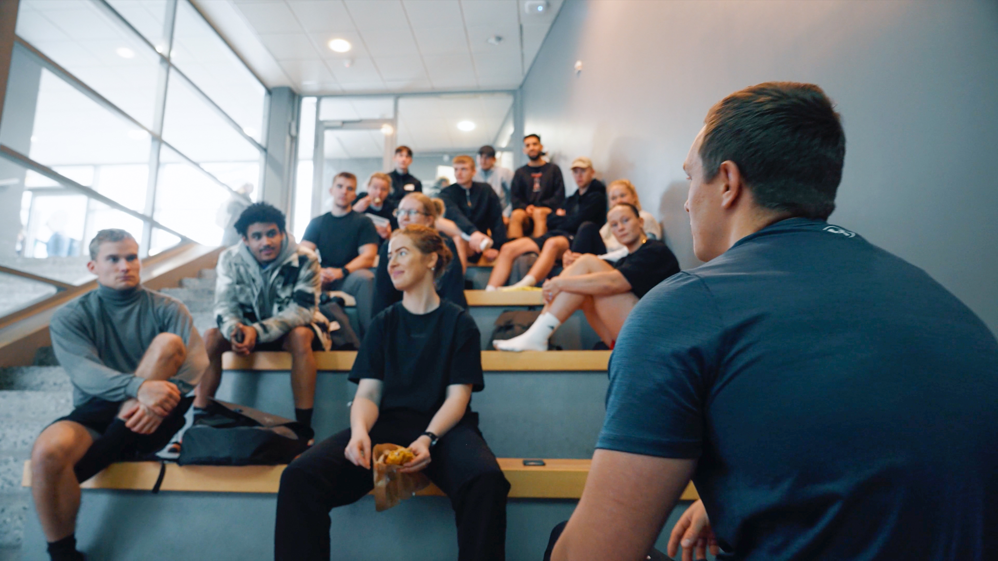 En gruppe unge voksne sidder på trappetrin i et moderne byggeri og lytter opmærksomt til en person, der taler foran dem. Stemningen er afslappet og fokuseret, og deltagerne er iført afslappet sportstøj. Nogle smiler og interagerer med hinanden.