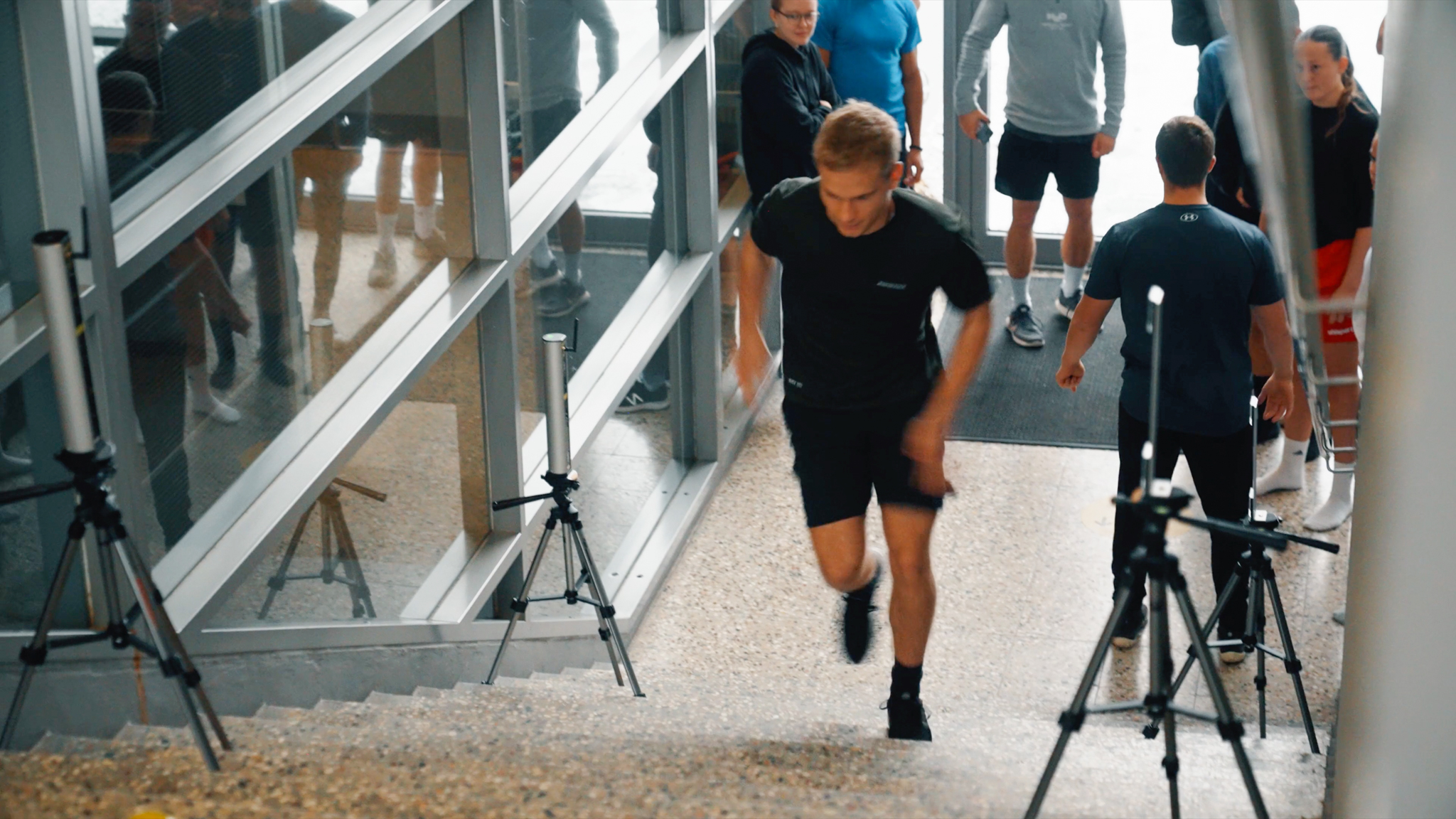 En mandlig atlet løber op ad en trappe i en bygning under en fysisk test, mens måleudstyr og sensorer på stativer er placeret langs trapperne. Flere personer står i baggrunden og observerer, muligvis som en del af et testhold eller træningsgruppe.