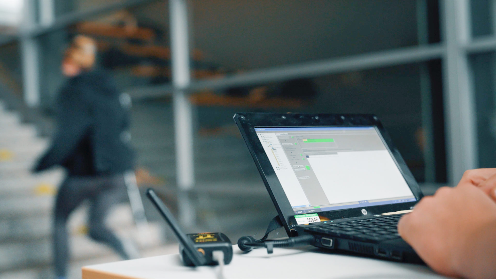 En person arbejder på en bærbar computer med måleudstyr tilsluttet, mens en atlet løber op ad en trappe i baggrunden. Skærmen viser et program til dataopsamling, sandsynligvis i forbindelse med en fysisk præstationstest. Miljøet ligner en sportshal eller et testcenter.