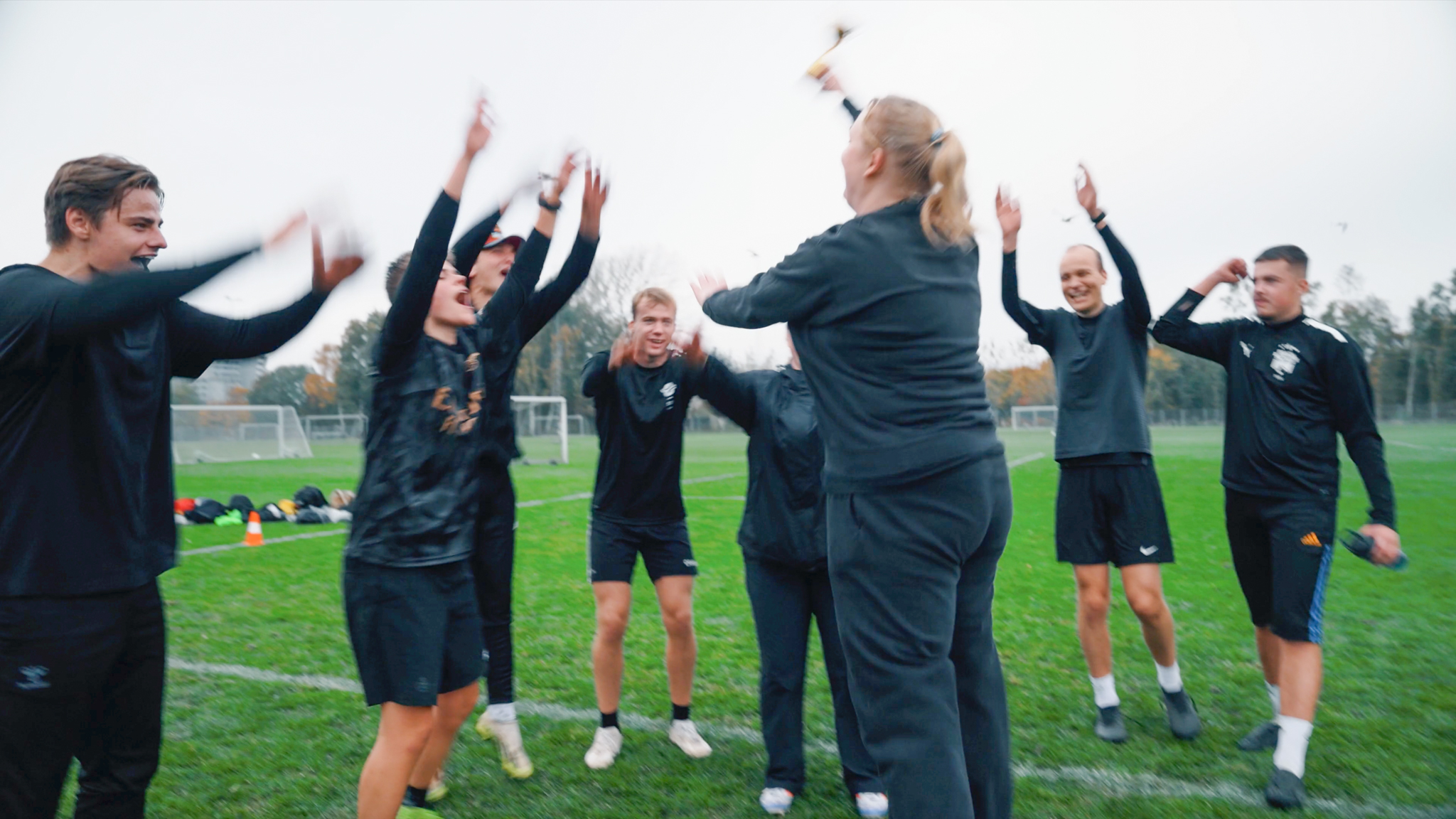 En gruppe unge i sportstøj fejrer sejren med jubel og hævede arme på en fodboldbane. En kvinde i midten holder en lille pokal i vejret, mens holdet omkring hende smiler og klapper. Det er overskyet, og der er mål og træningsudstyr i baggrunden.