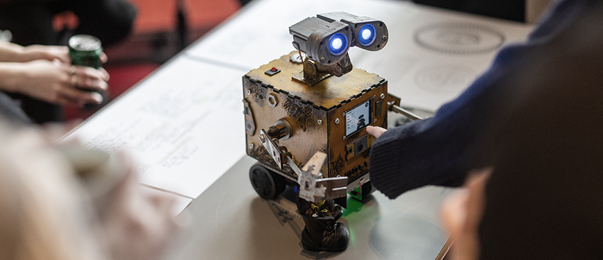 Selvbygget, firkantet robot af laserskåret krydsfinér med 2 øjne og hjul, der står på et projektbord, og to par hænder, hvoraf det ene par justere noget på robotten.
