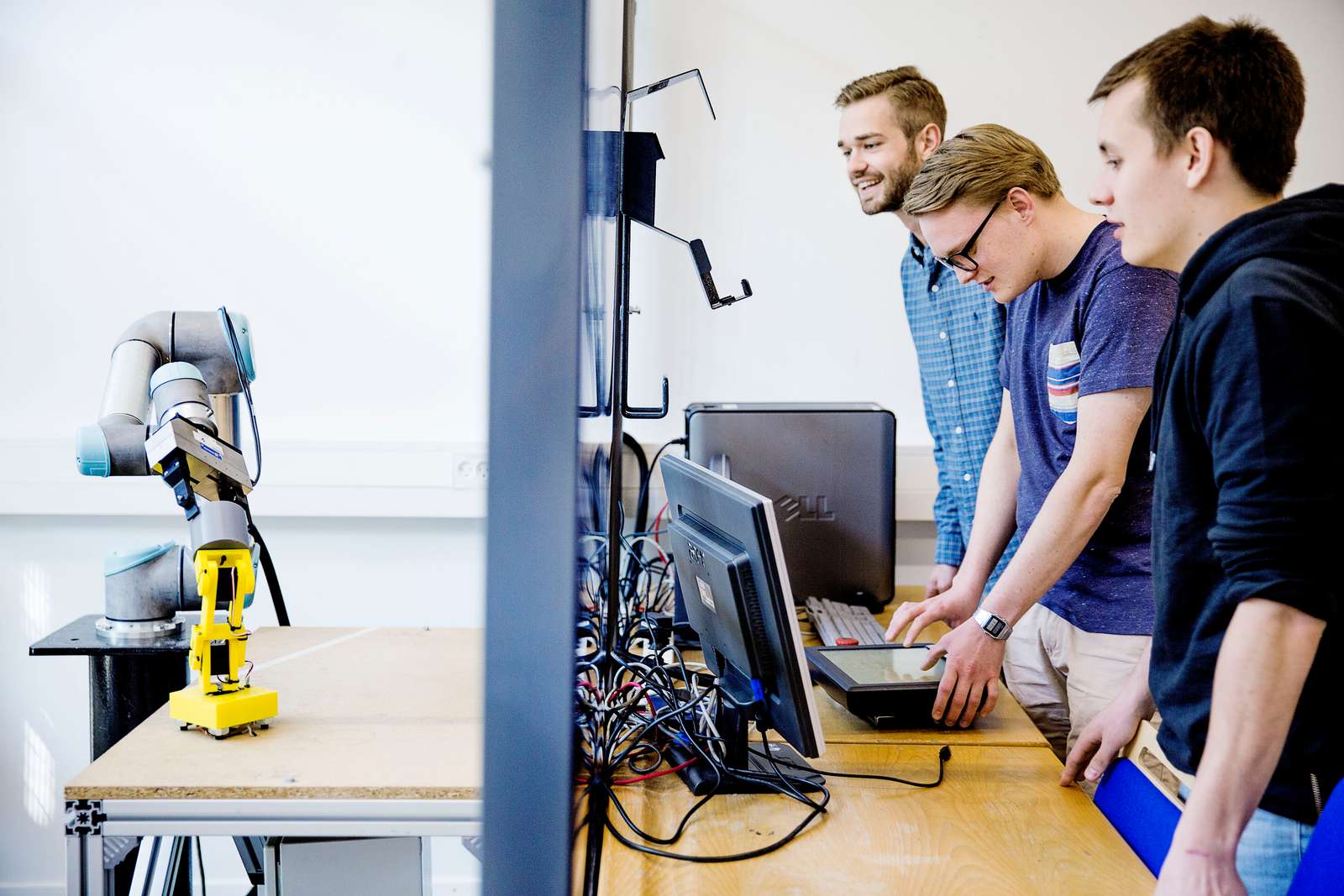 Tre studerende observerer og betjener computere ved siden af en robotarm, som flytter en gul enhed på et bord i et laboratorium.