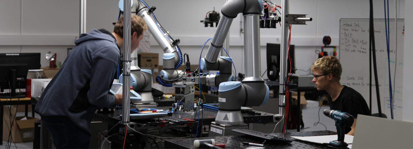 To personer arbejder ved en robotcelle med flere robotarme og teknisk udstyr i et laboratoriemiljø.