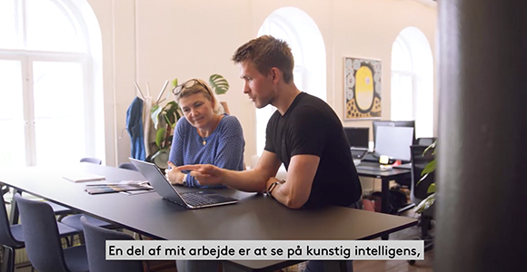 To personer samarbejder ved en bærbar computer på et kontor med teksten "En del af mit arbejde er at se på kunstig intelligens," nederst i billedet.