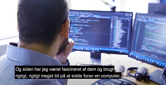 Person set bagfra foran to skærme med kode og underteksten "Og siden har jeg været fascineret af dem og brugt rigtigt, rigtigt meget tid på at sidde foran en computer,"