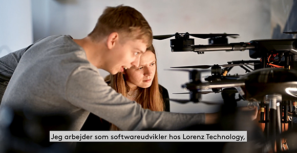 To personer undersøger og justerer en stor drone indendørs, med underteksten "Jeg arbejder som softwareudvikler hos Lorenz Technology," nederst i billedet.