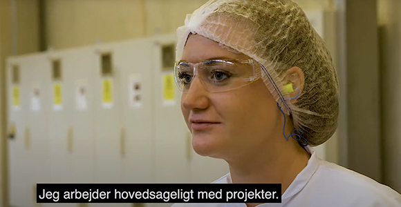 Kvinde i laboratoriebeklædning med hårnet og beskyttelsesbriller i et industrielt miljø med underteksten "Jeg arbejder hovedsageligt med projekter."
