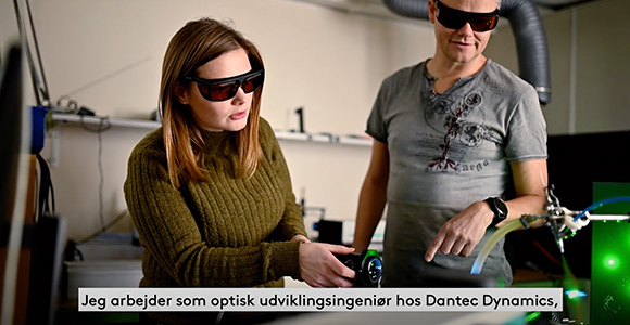 To personer med mørke beskyttelsesbriller arbejder ved laboratorieudstyr. Teksten nederst siger: "Jeg arbejder som optisk udviklingsingeniør hos Dantec Dynamics,"