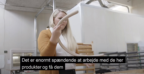 Kvinde i et værksted holder et bøjet emne med teksten "Det er enormt spændende at arbejde med de her produkter og få dem" nederst i billedet.