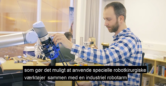 Mand betjener en industriel robotarm i et laboratoriemiljø med underteksten "som gør det muligt at anvende specielle robotkirurgiske værktøjer sammen med en industriel robotarm".