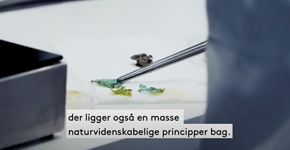 Nærbillede af laboratoriearbejde med et redskab og små prøver på en overflade samt teksten "der ligger også en masse naturvidenskabelige principper bag."