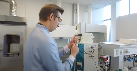 Person i laboratoriekittel arbejder med laboratorieudstyr i et lyst kemi- eller bioteknologilaboratorium.